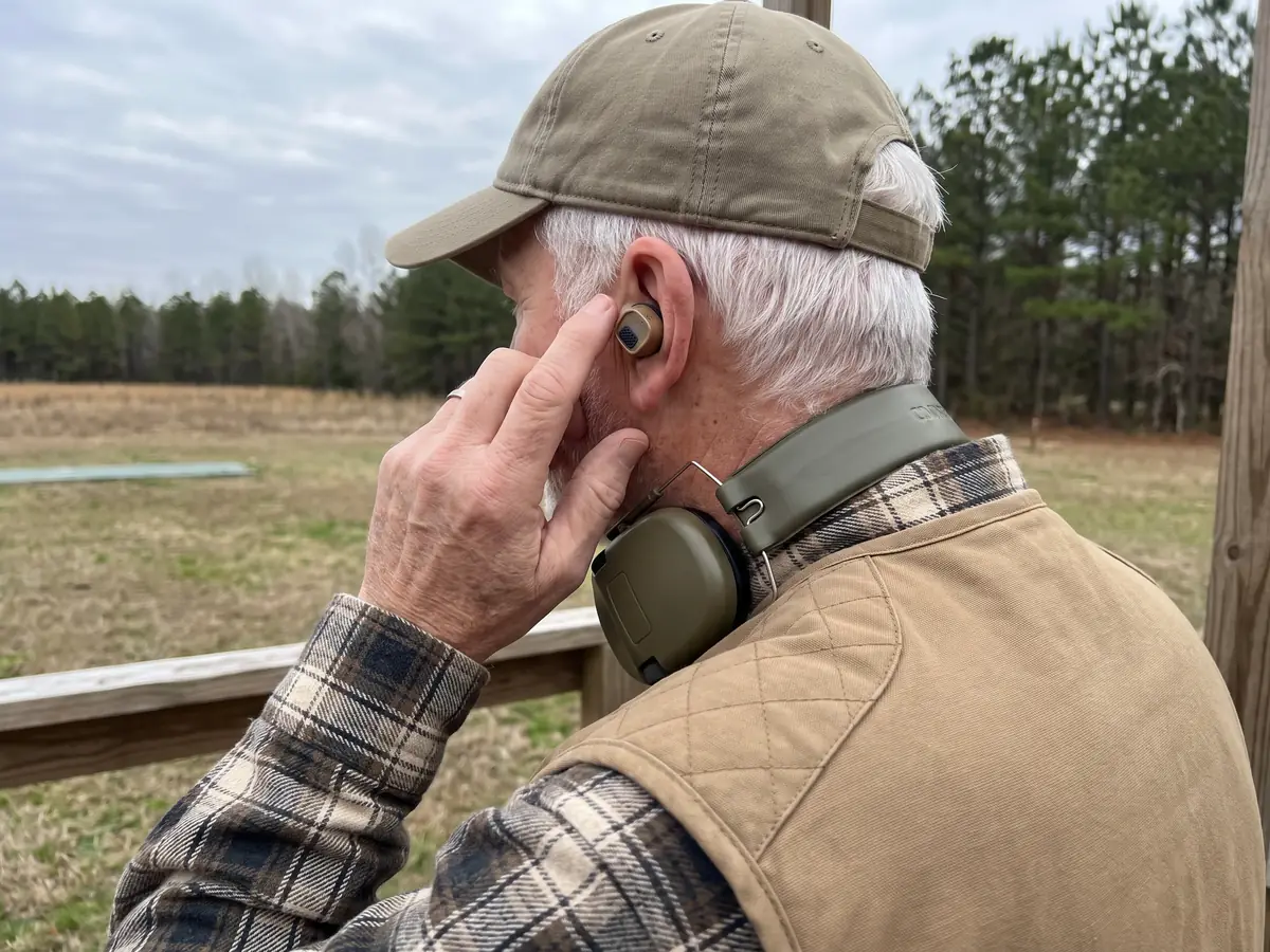 An older man outdoors pressing a FieldOps Pro earbud into his ear, a pair of ear muffs round his neck